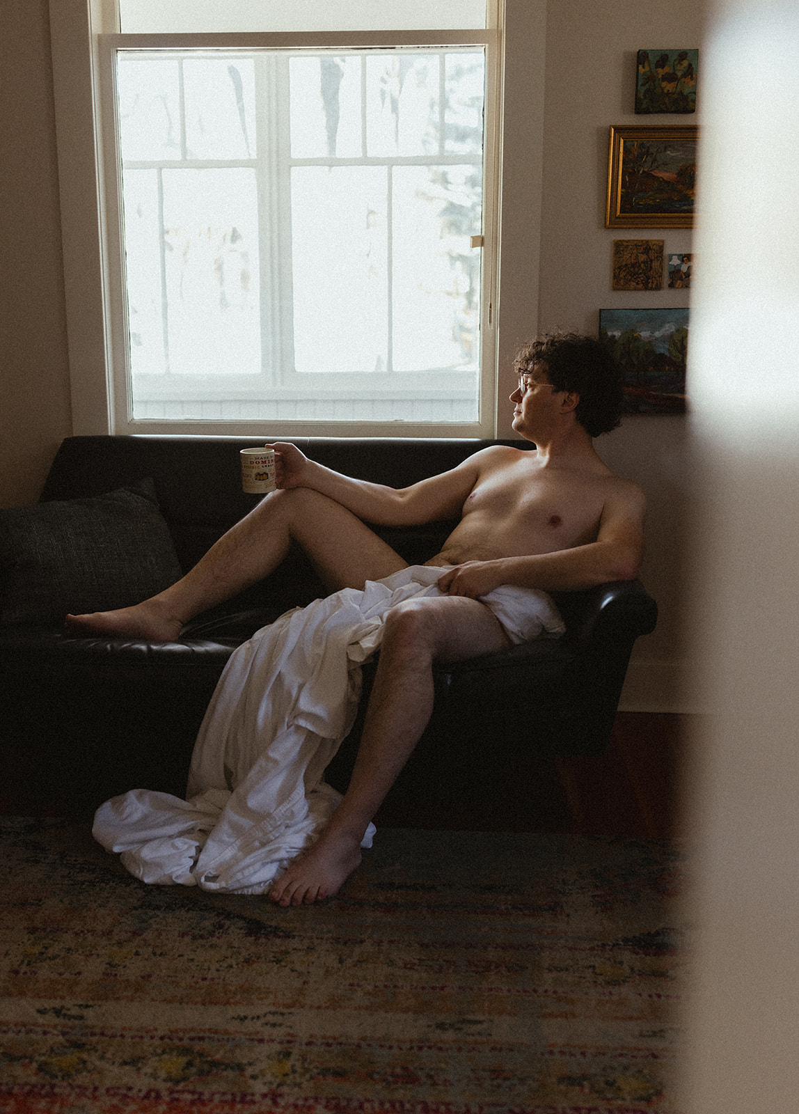 naked man with curly hair sitting on a couch by a window with a white sheet covering his body
