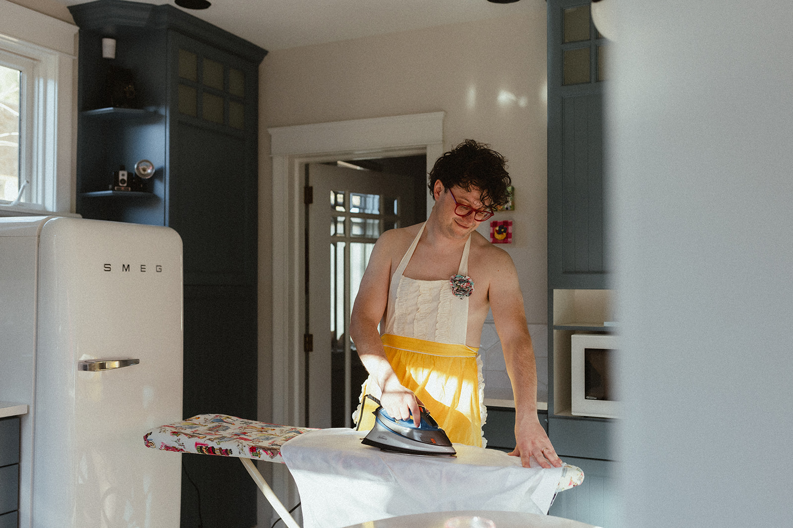 young naked man with nothing but an apron on while ironing a white button-down shirt